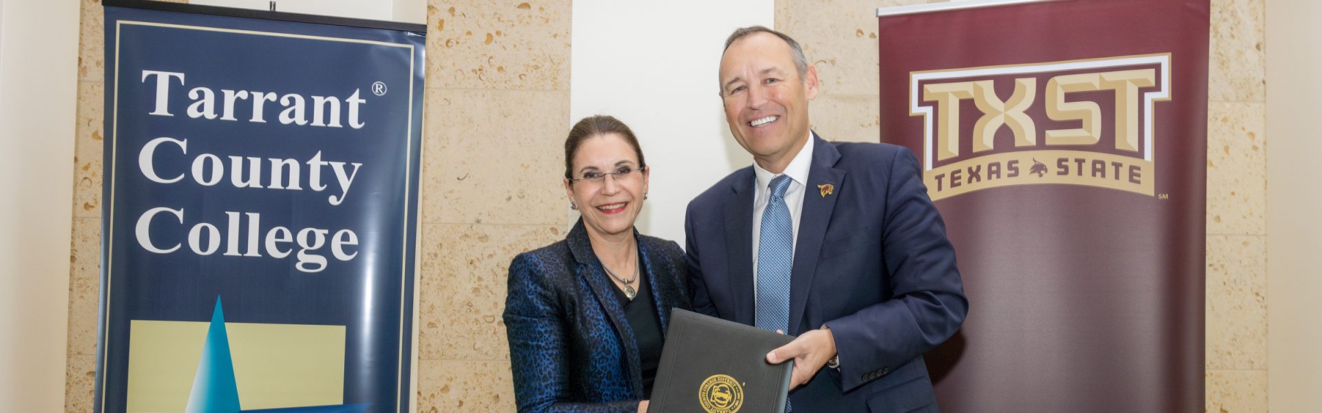 TCC Chancellor Elva LeBlanc and Texas State President Kelly Damphousse hold a folio containing the agreement documents between them and pose in front of banners for the two instituitons